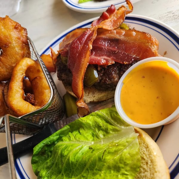 A bacon cheeseburger with a side of onion rings, lettuce, and dipping sauce on a plate.